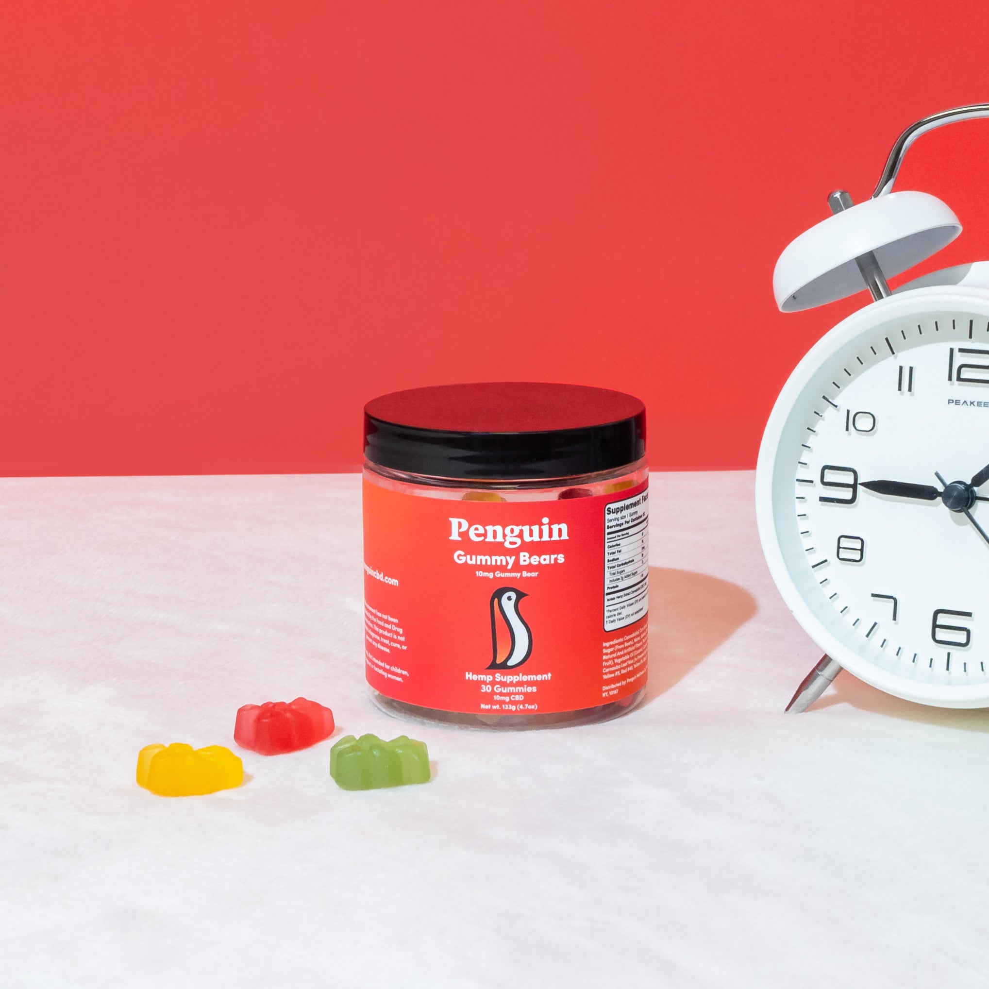 Jar of Penguin Gummy Bears with colorful gummy bears and a white alarm clock on a red and white background.